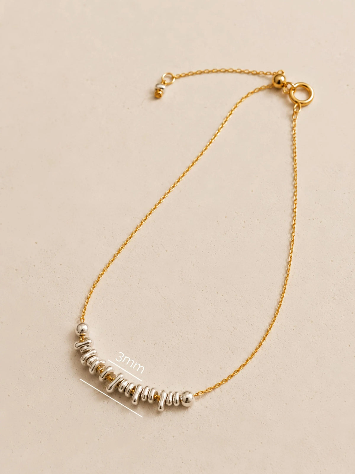 A mixed metal bracelet with delicate organic shaped sterling silver beads on a fine gold chain with measurement label on a beige background