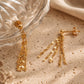 A pair of 18k gold-plated sterling silver tassel earrings draped over a textured glass bowl and a beige stone, showcasing the shimmering, light-catching flat-link fringe chains.