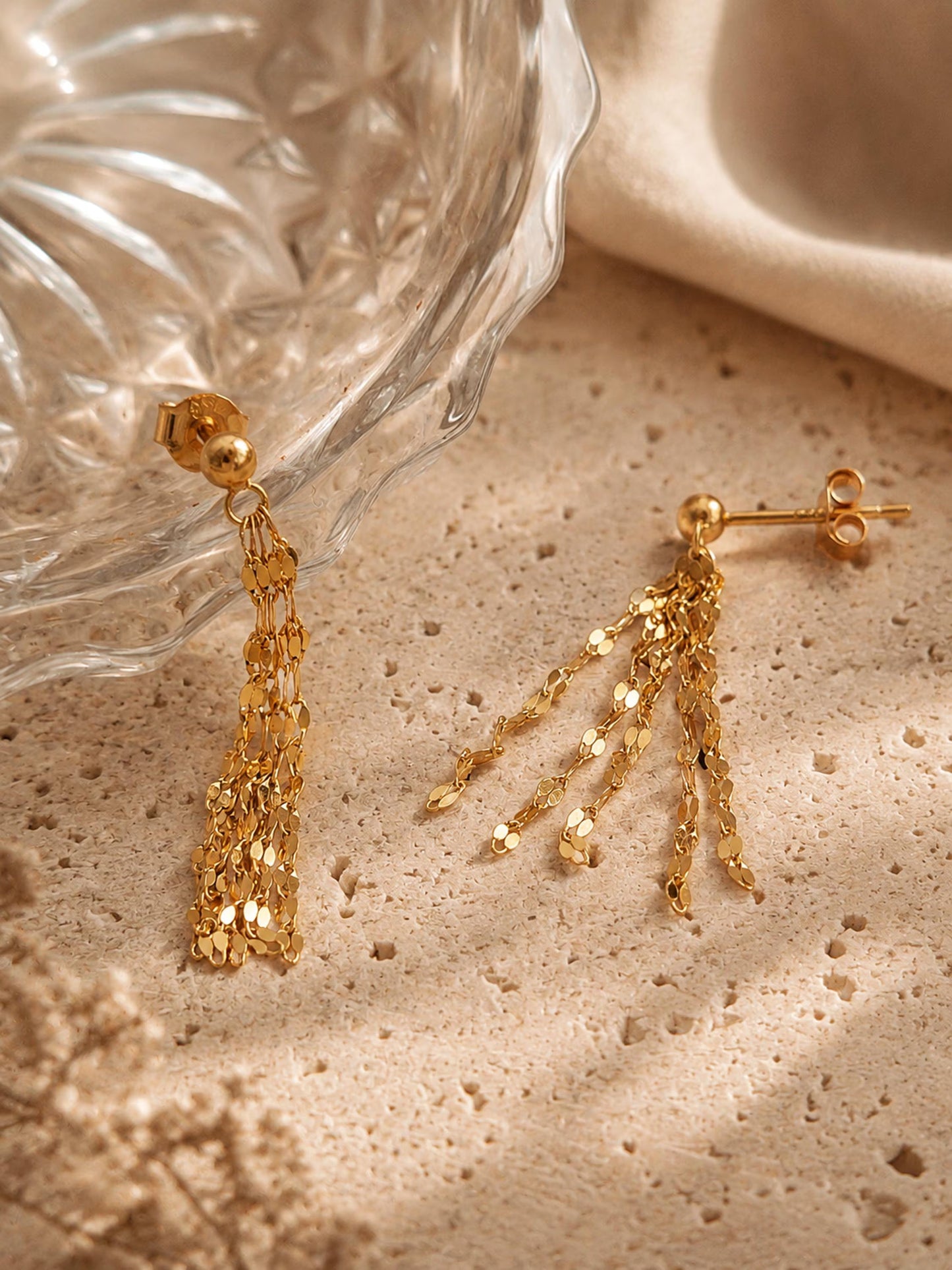 A pair of 18k gold-plated sterling silver tassel earrings draped over a textured glass bowl and a beige stone, showcasing the shimmering, light-catching flat-link fringe chains.