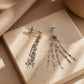 A pair of sterling silver tassel earrings resting on a neutral beige stone tile, displaying the delicate flat-link fringe chains draped straight and fanned out in warm, soft sunlight.