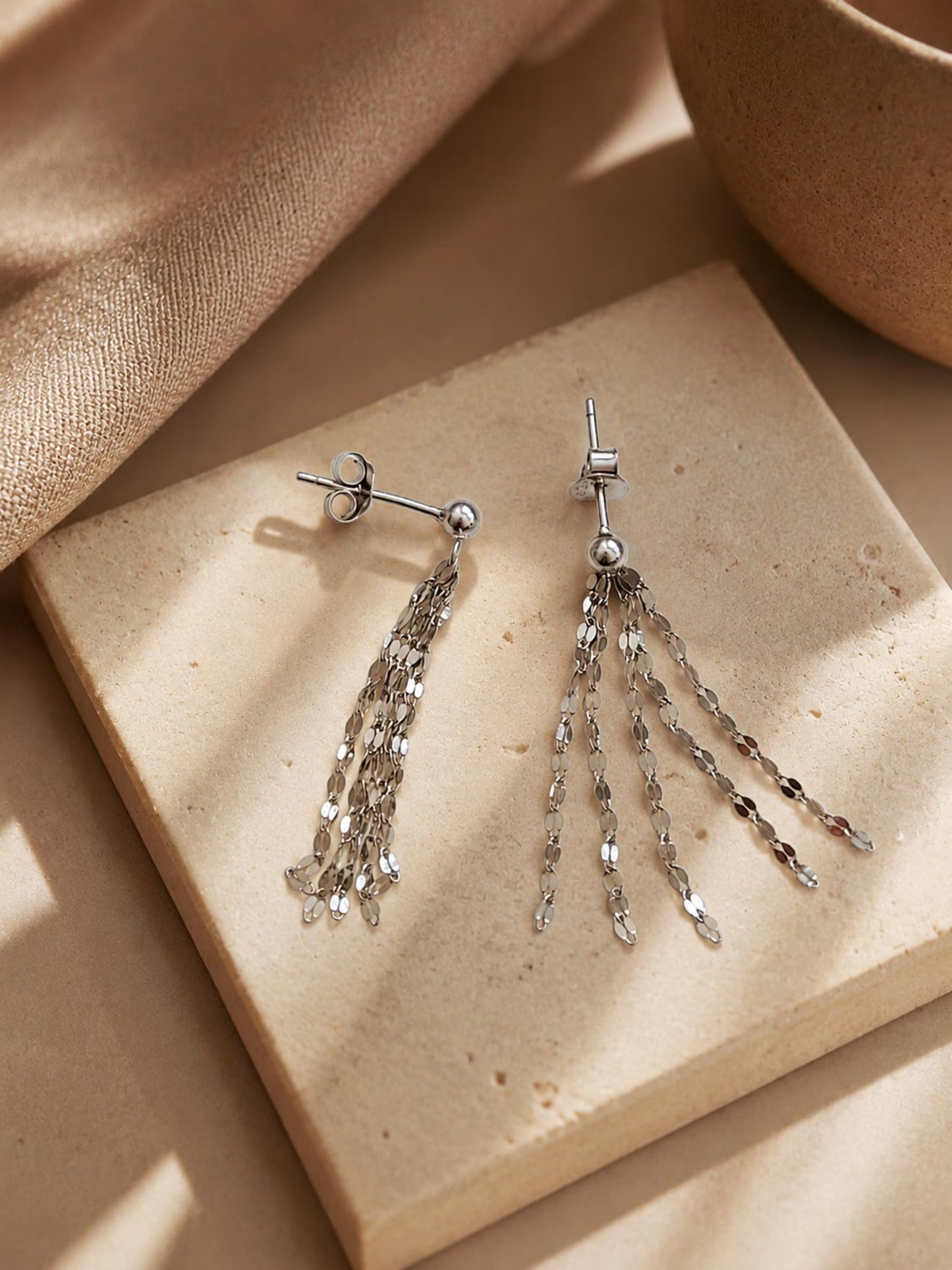 A pair of sterling silver tassel earrings resting on a neutral beige stone tile, displaying the delicate flat-link fringe chains draped straight and fanned out in warm, soft sunlight.
