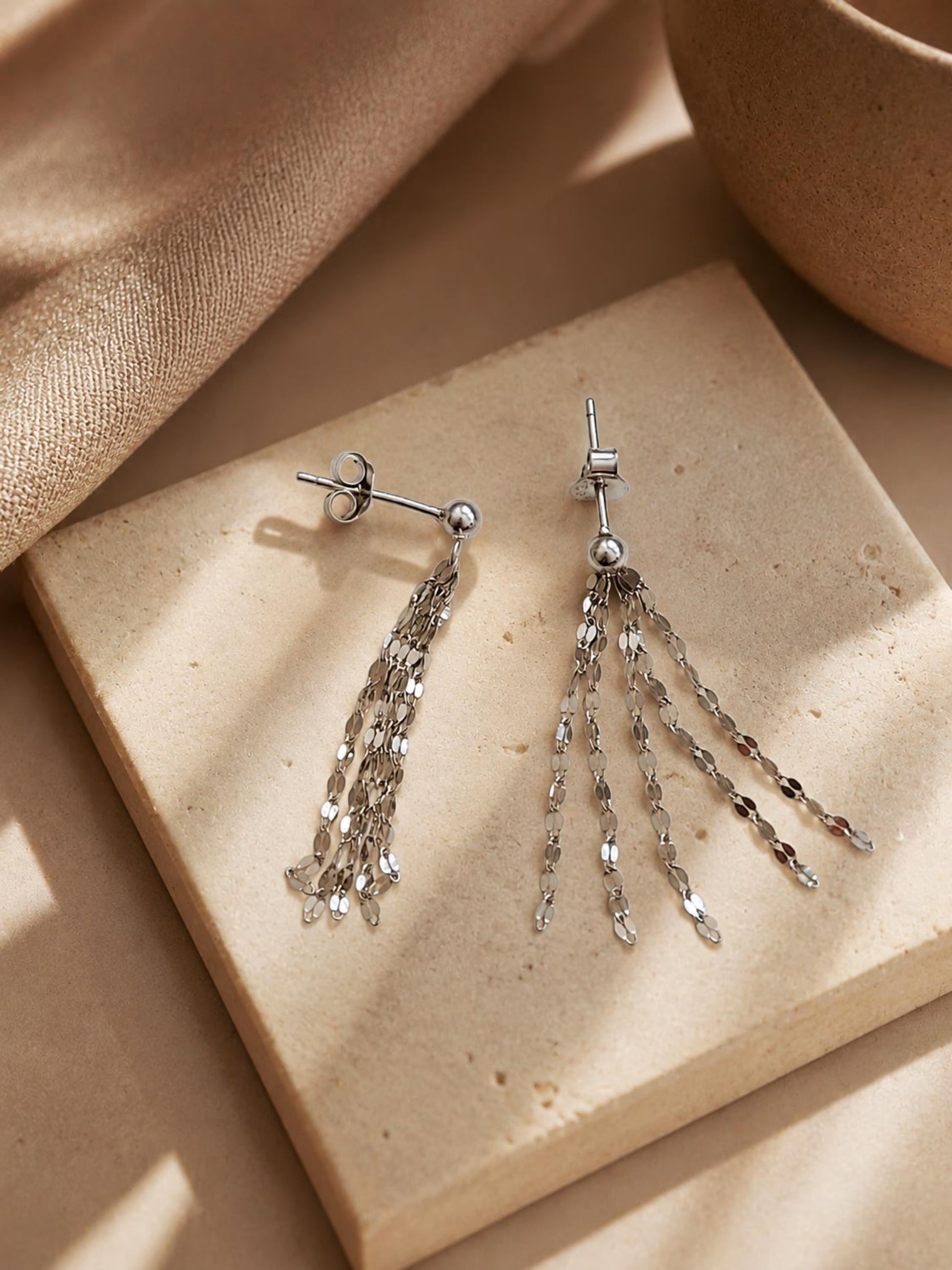 A pair of sterling silver tassel earrings resting on a neutral beige stone tile, displaying the delicate flat-link fringe chains draped straight and fanned out in warm, soft sunlight.