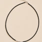 A necklace featuring a strand of small, faceted black spinel beads arranged in a circular shape. A short section of silver beads is positioned at the bottom centre. The necklace includes a silver-toned chain extender with a small circular tag.