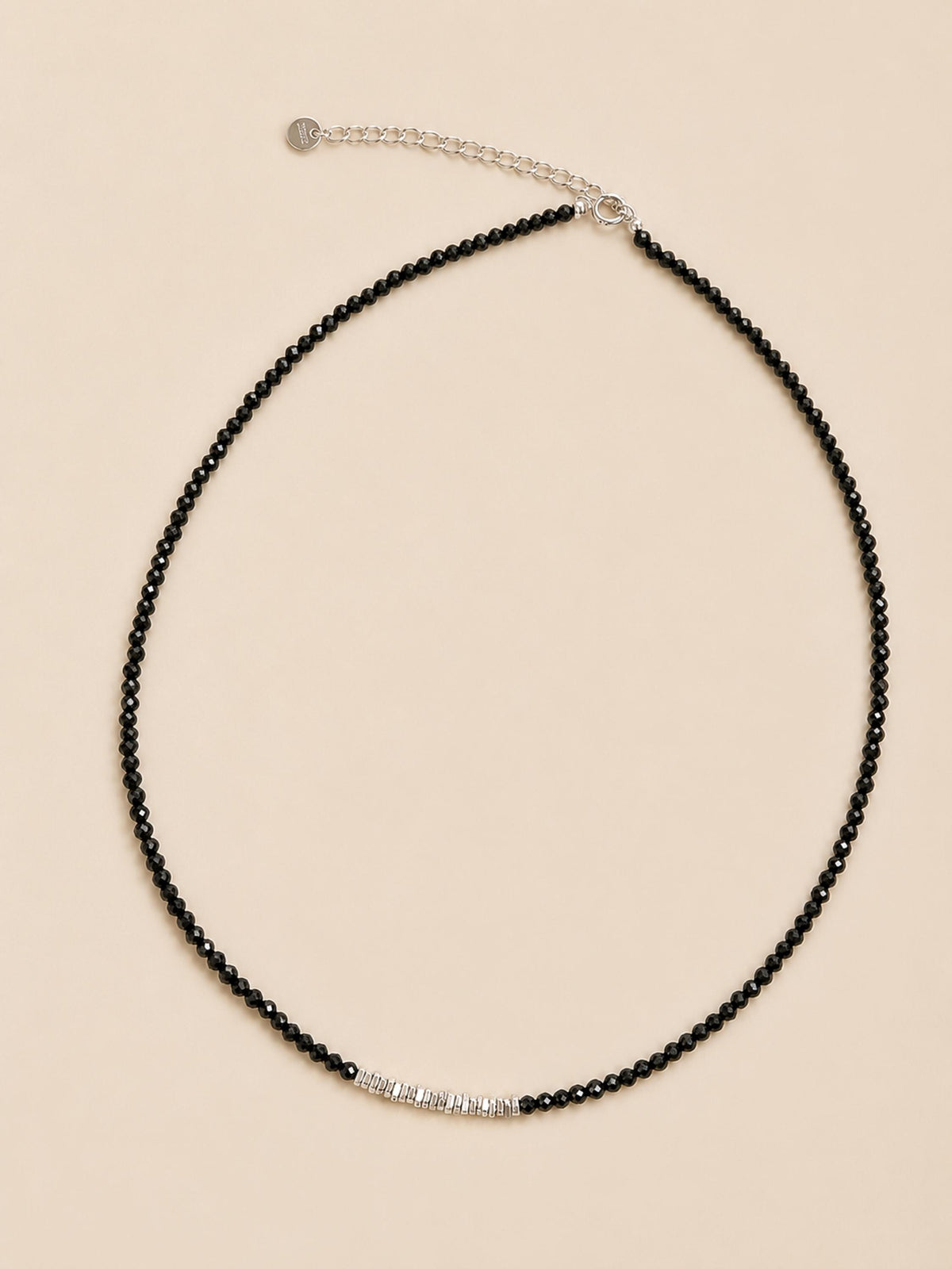 A necklace featuring a strand of small, faceted black spinel beads arranged in a circular shape. A short section of silver beads is positioned at the bottom centre. The necklace includes a silver-toned chain extender with a small circular tag.
