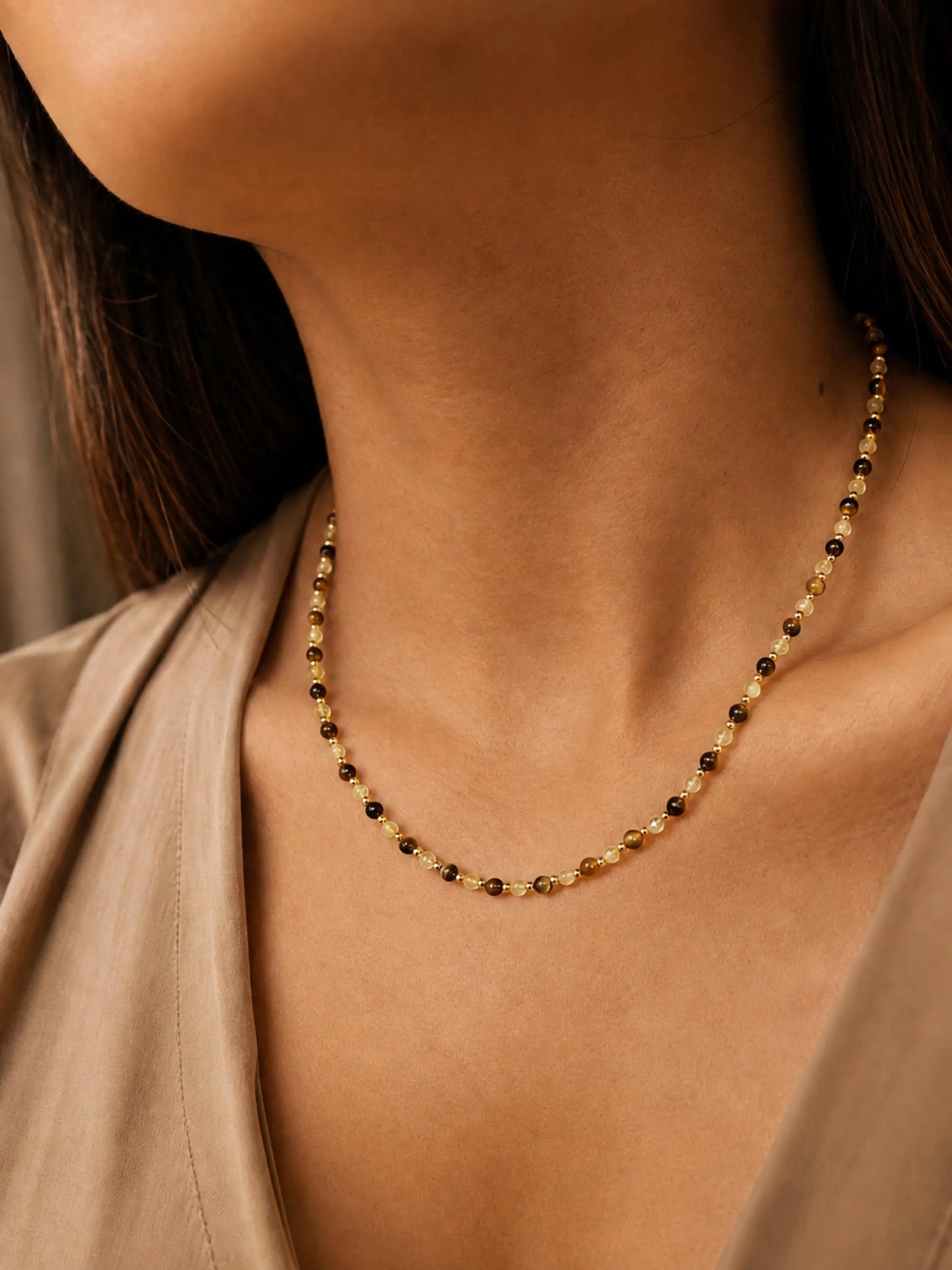 Close-up model photo showcasing a delicate, dainty beaded necklace made of warm brown tiger's eye and light yellow citrine beads. The necklace sits gracefully on the collarbone, styled against a soft beige v-neck top.