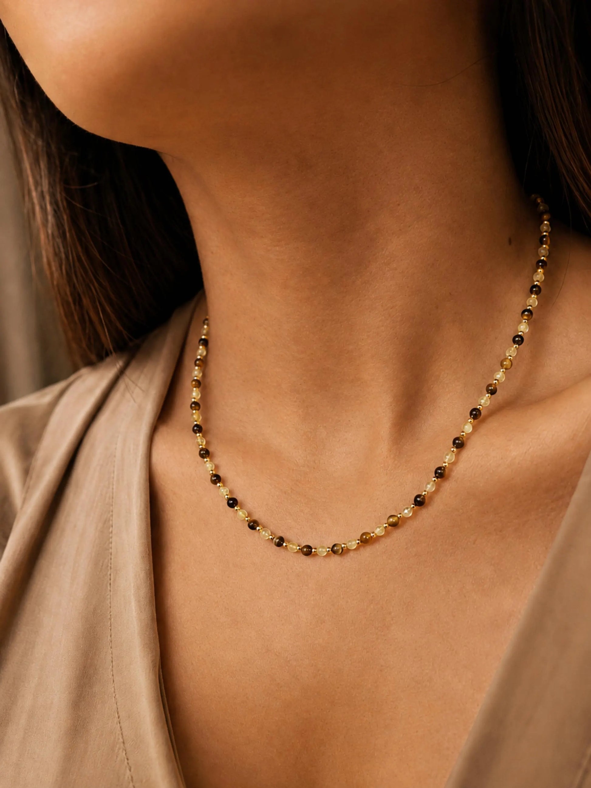 Close-up model photo showcasing a delicate, dainty beaded necklace made of warm brown tiger's eye and light yellow citrine beads. The necklace sits gracefully on the collarbone, styled against a soft beige v-neck top.