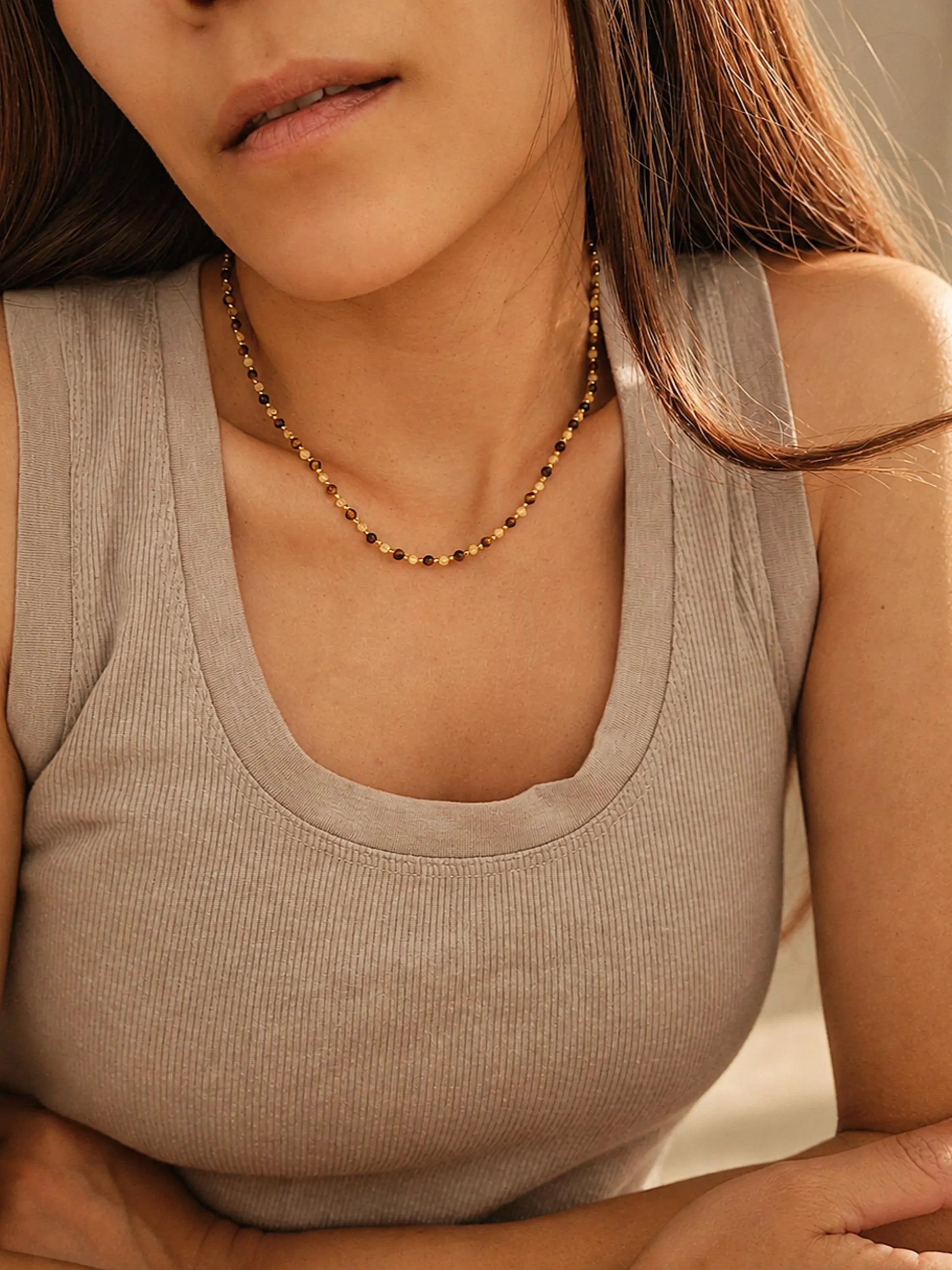 Close-up model photo showcasing a delicate, dainty beaded necklace made of warm brown tiger's eye and light yellow citrine beads. The necklace sits gracefully on the collarbone, styled against a soft beige tank top.