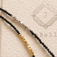 May include: Two beaded necklaces featuring small, faceted black beads. One necklace includes a section of silver-toned metallic nuggets, while the other features a section of gold-toned metallic nuggets. The necklaces are displayed against a textured surface.