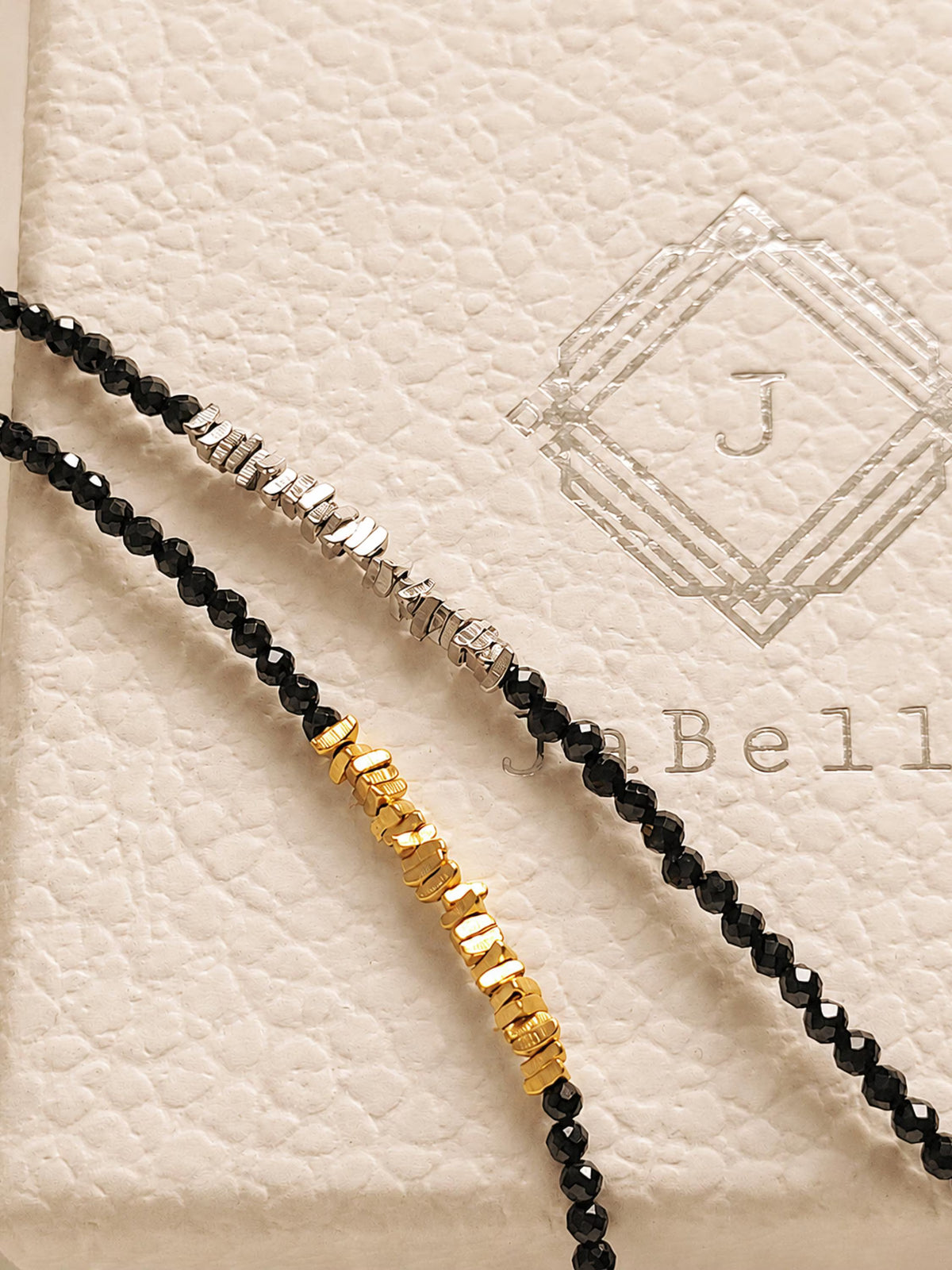 May include: Two beaded necklaces featuring small, faceted black beads. One necklace includes a section of silver-toned metallic nuggets, while the other features a section of gold-toned metallic nuggets. The necklaces are displayed against a textured surface.