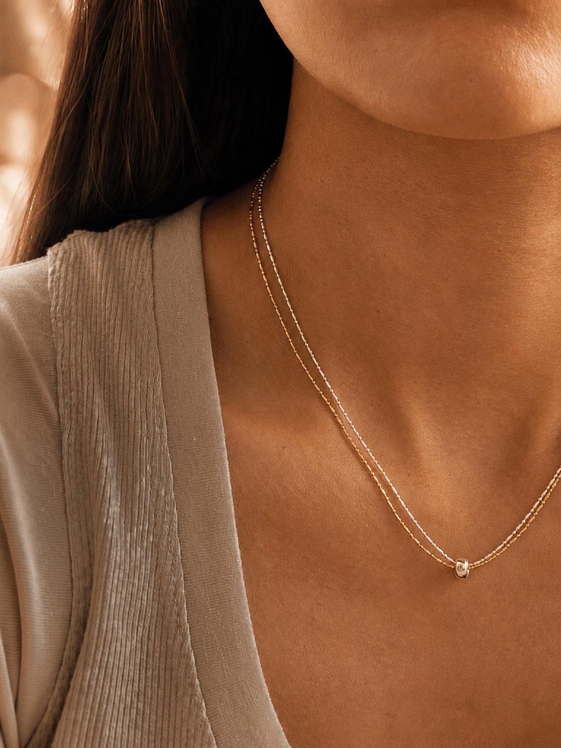 A close-up lifestyle shot of a model wearing a delicate double-strand gold necklace with a small pendant, styled with a beige ribbed top.