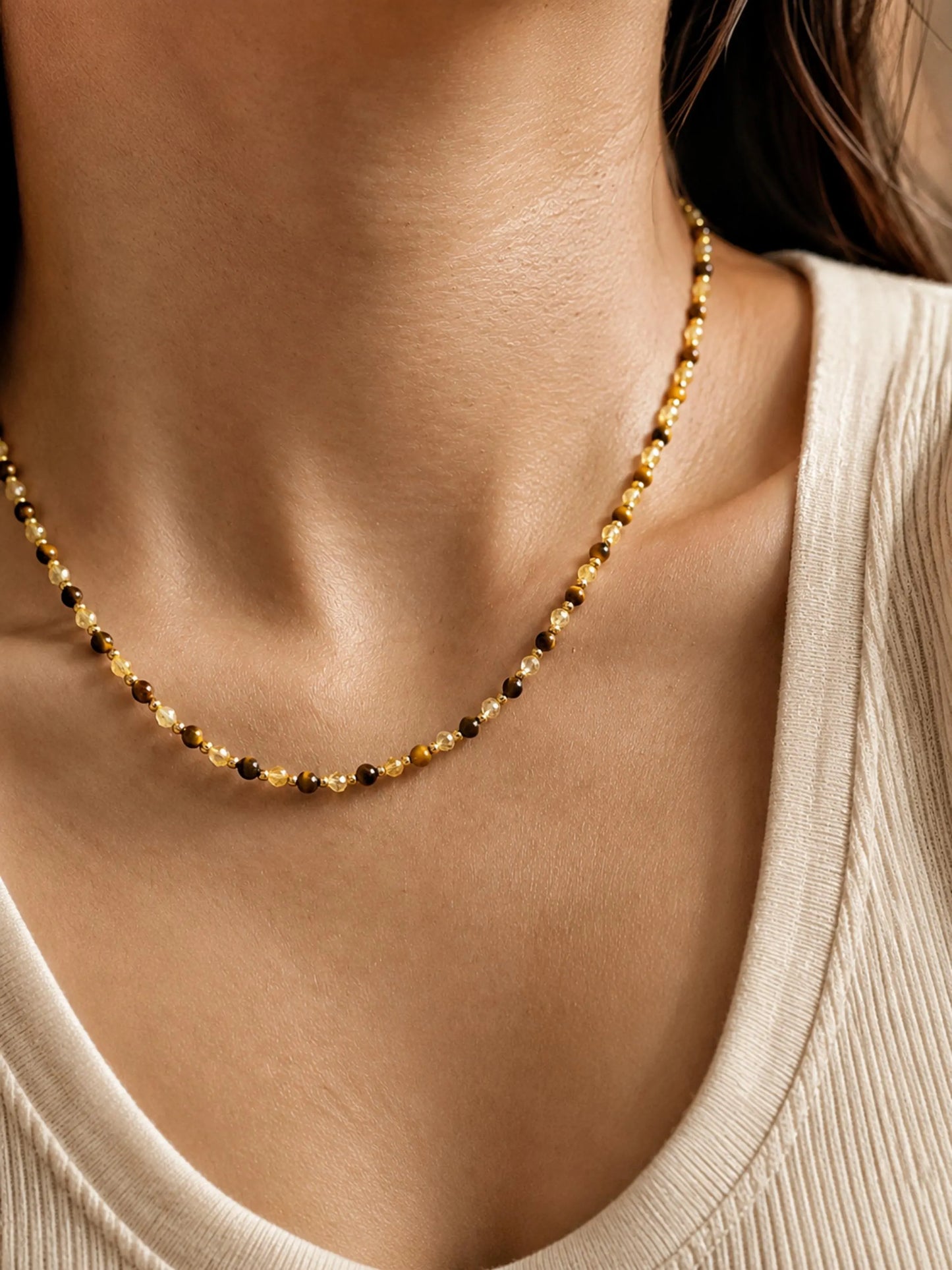 Close-up model photo showcasing a delicate, dainty beaded necklace made of warm brown tiger's eye and light yellow citrine beads. The necklace sits gracefully on the collarbone, styled against a soft beige tank top.