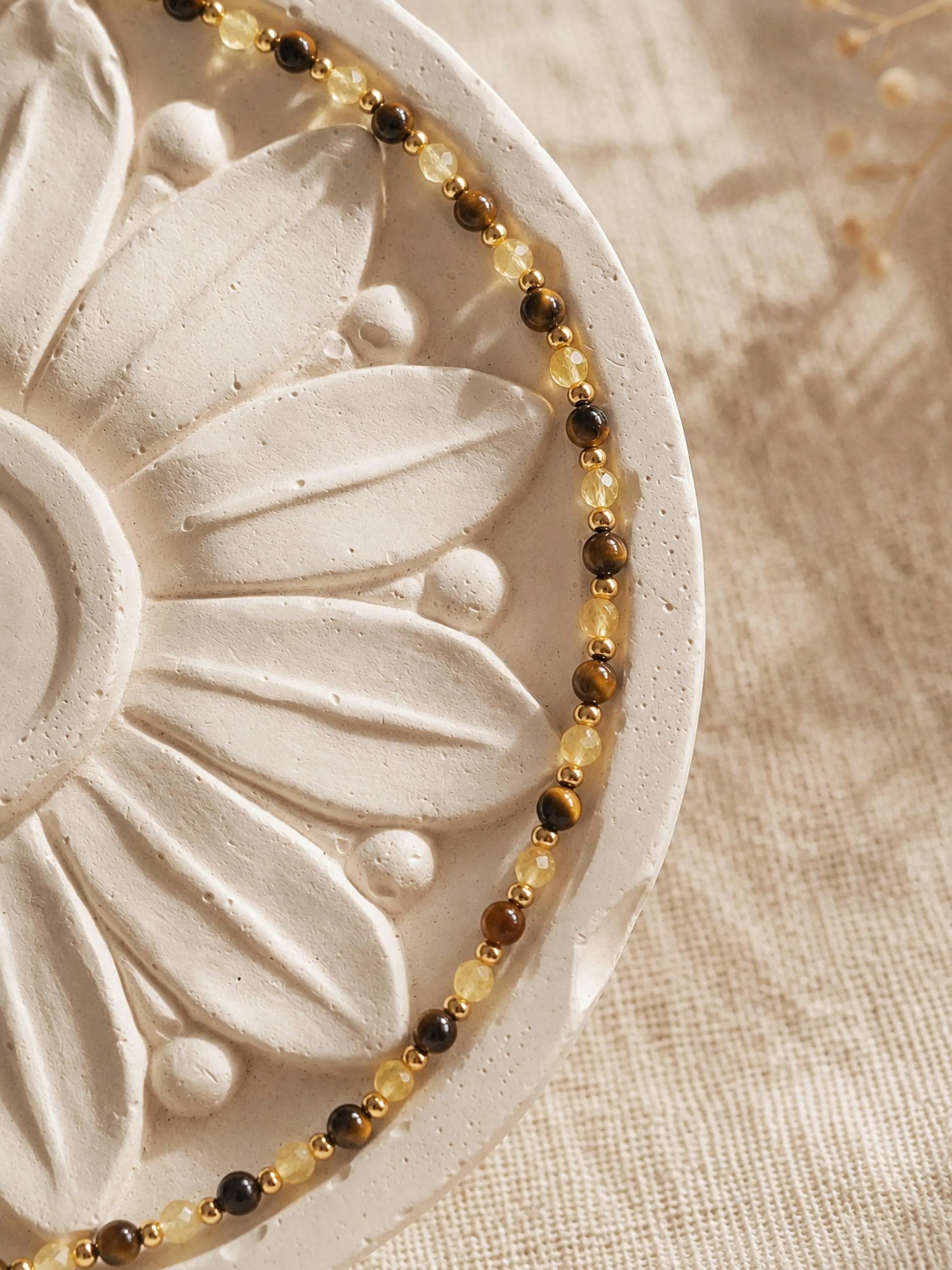 A delicate, dainty beaded necklace made of warm brown tiger's eye and light yellow citrine beads with gold accents displayed on a luxury flower plate.