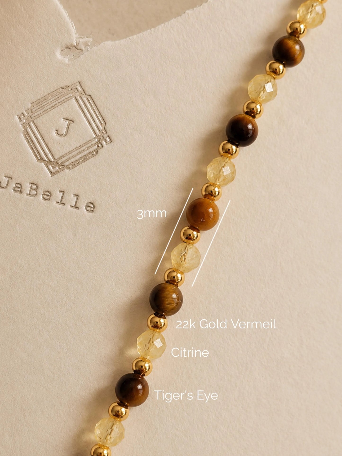 Close-up of a delicate gemstone necklace featuring alternating smooth round tiger's eye beads, faceted translucent yellow citrine beads, and small 22k gold vermeil spacers. Annotations show 3mm bead sizing set against a warm beige textured background with an embossed JaBelle Jewellery logo.