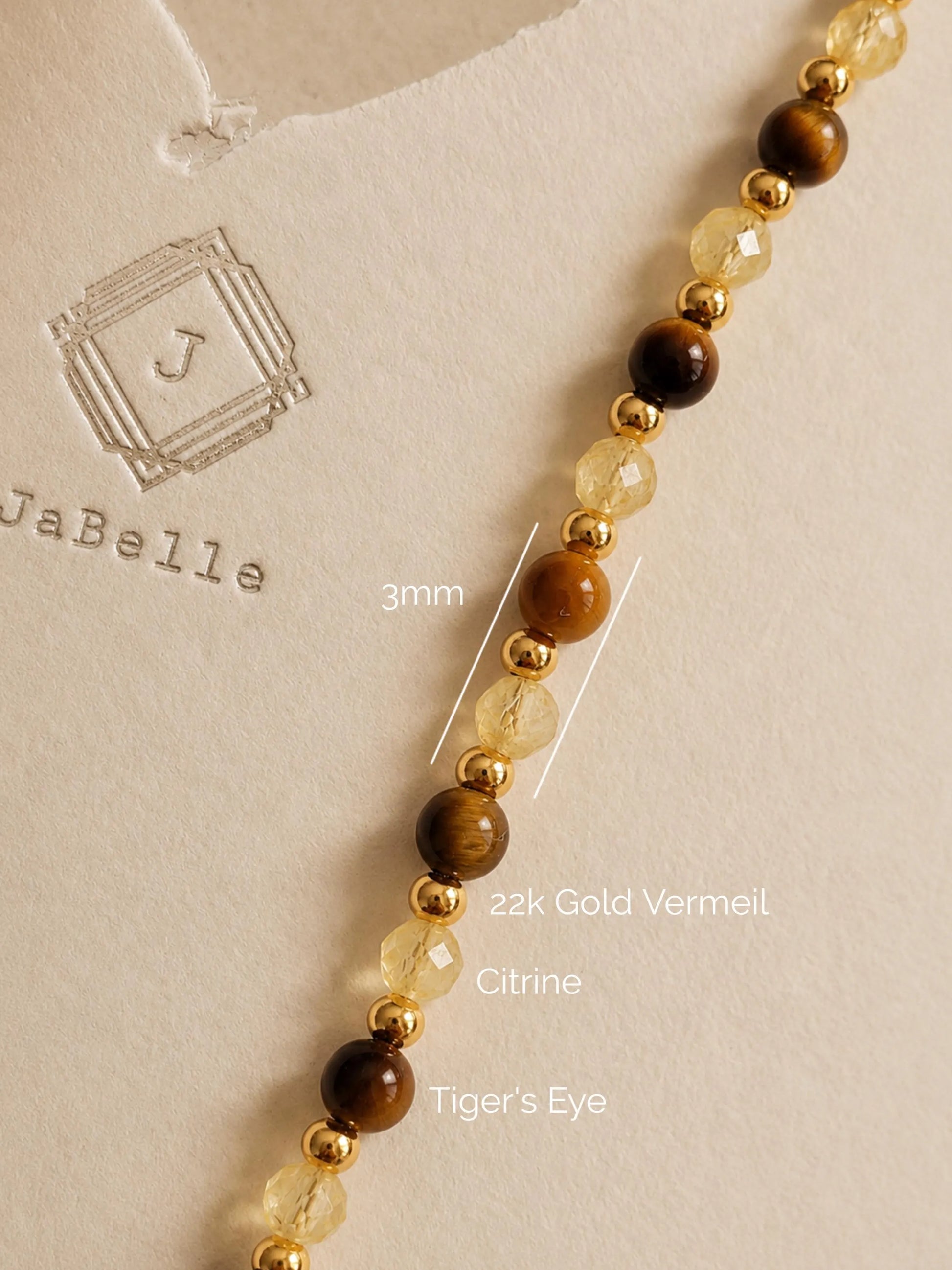Close-up of a delicate gemstone necklace featuring alternating smooth round tiger's eye beads, faceted translucent yellow citrine beads, and small 22k gold vermeil spacers. Annotations show 3mm bead sizing set against a warm beige textured background with an embossed JaBelle Jewellery logo.
