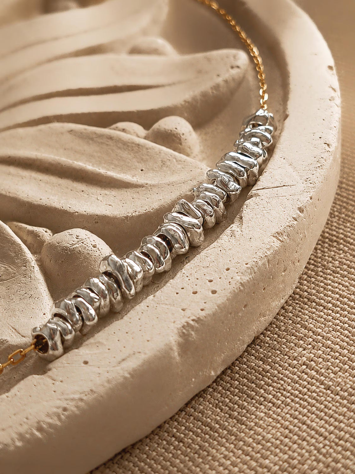 Detailed view of sculptural sterling silver nugget beads arranged along a gold chain on a curved beige stone surface.