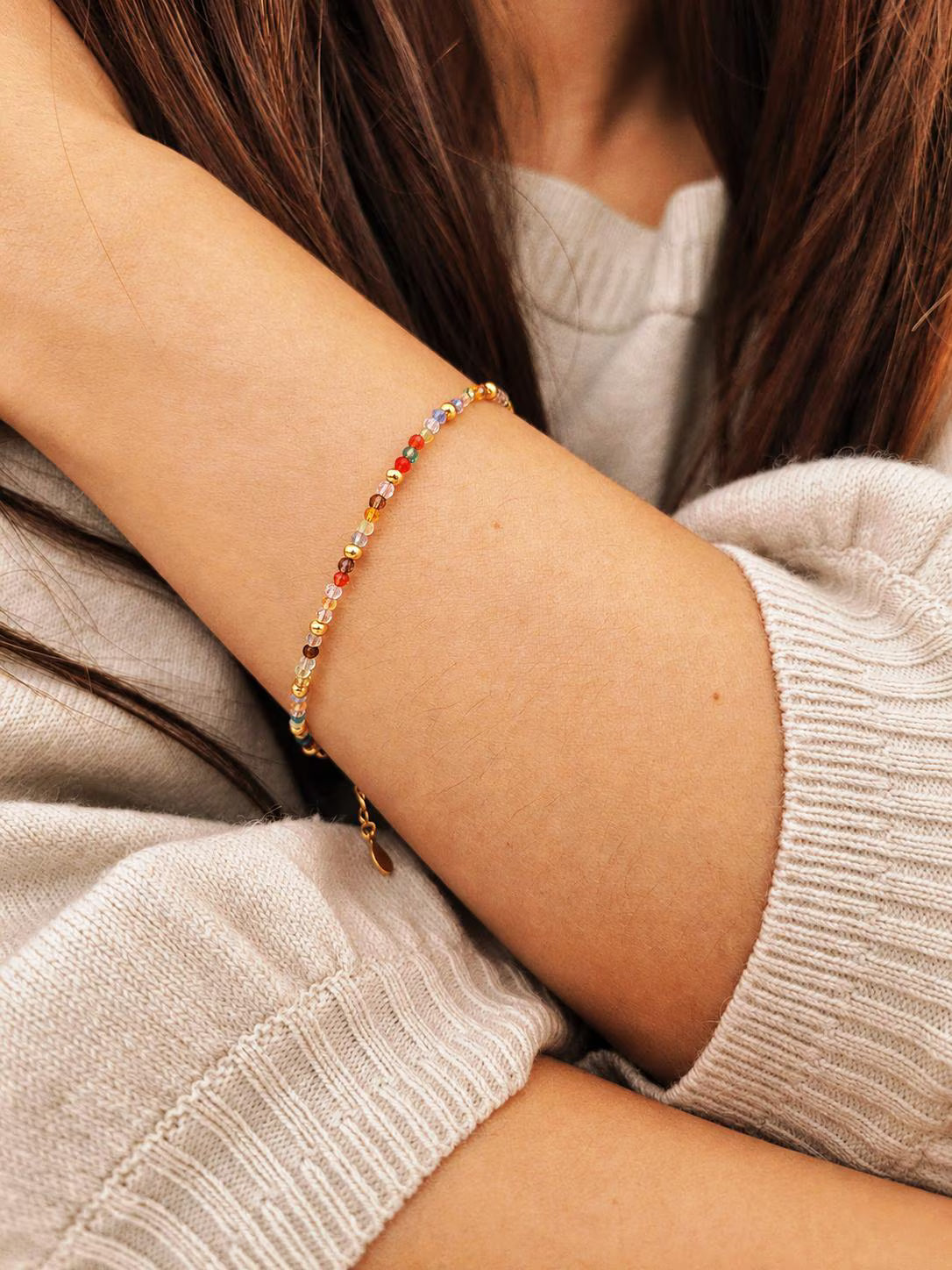 A delicate gold chain bracelet with small colorful crystal beads worn on a girl's wrist, shown in a close-up lifestyle shot with a ribbed beige sweater.