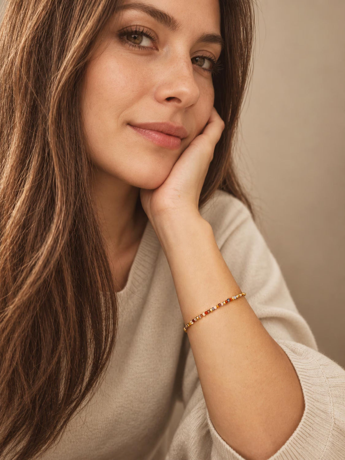 A woman with long brown hair rests her head on her hand, wearing a delicate gold beaded bracelet with small red and orange beads. She is wearing a light beige knit sweater against a soft, warm beige background.