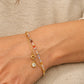 A close-up of a lady's wrist wearing a delicate gold bracelet with a small clear stone charm and a thin crystal beaded chain with small multicolored beads, set against a beige knit top.