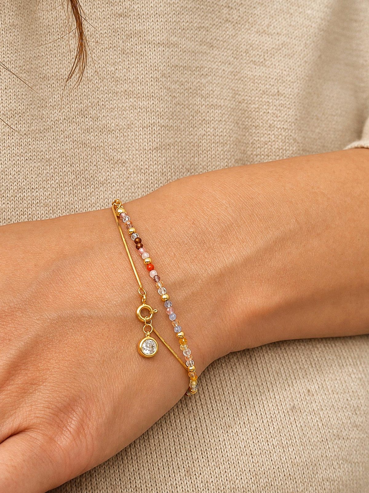 A close-up of a lady's wrist wearing a delicate gold bracelet with a small clear stone charm and a thin crystal beaded chain with small multicolored beads, set against a beige knit top.