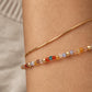 A close-up of a woman's wrist wearing two delicate bracelets: a fine gold snake chain and a beaded bracelet with faceted multicolor crystal beads in orange, red, blue, and gold.