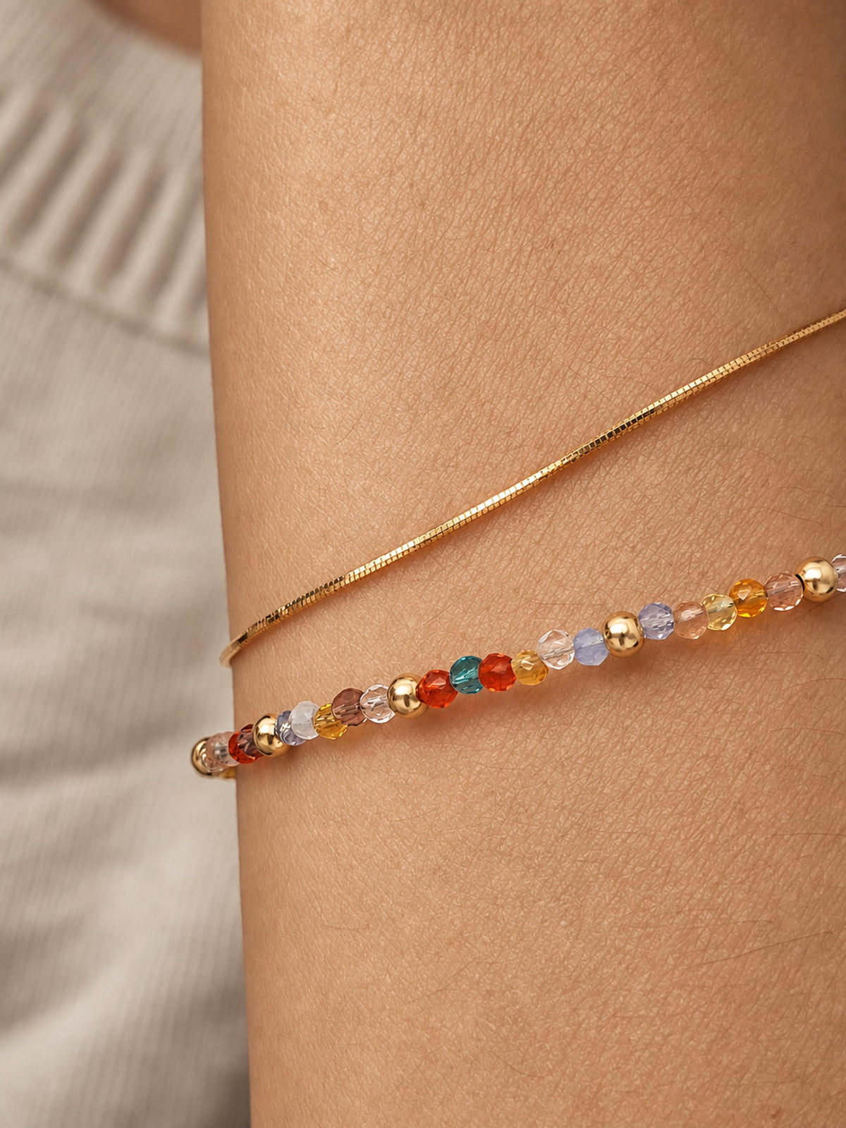 A close-up of a woman's wrist wearing two delicate bracelets: a fine gold snake chain and a beaded bracelet with faceted multicolor crystal beads in orange, red, blue, and gold.