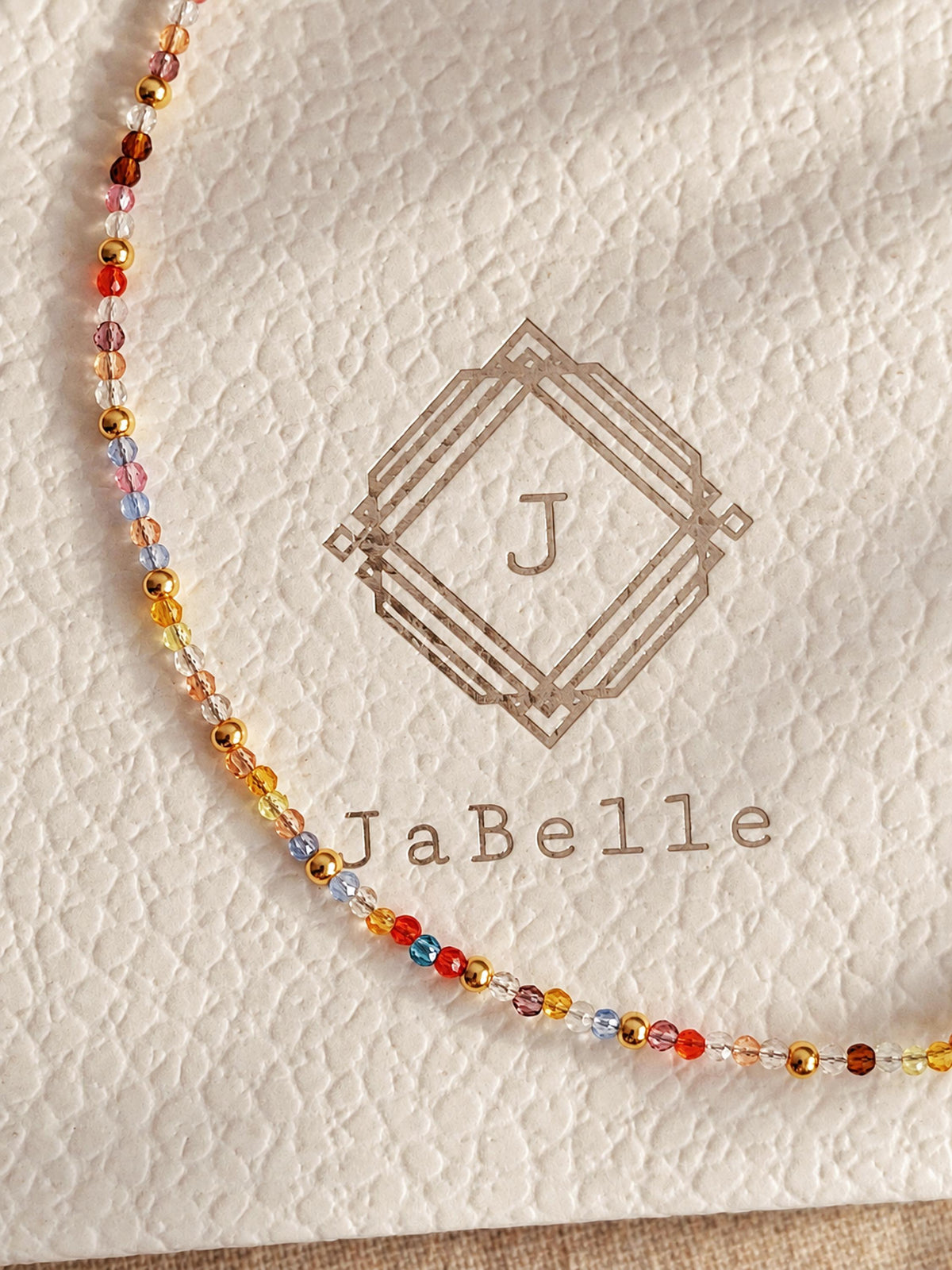 A close-up of a delicate beaded strand with small multicolored crystal beads, laid on textured cream packaging printed with the brand name Jabelle.