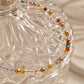 A delicate crystal beaded bracelet with gold and multicolor faceted beads draped over a clear textured glass jar on a soft beige fabric background.