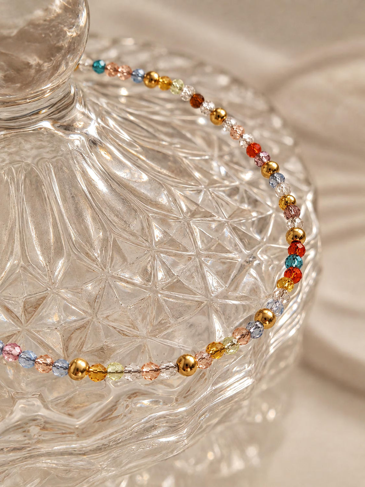 A delicate crystal beaded bracelet with gold and multicolor faceted beads draped over a clear textured glass jar on a soft beige fabric background.