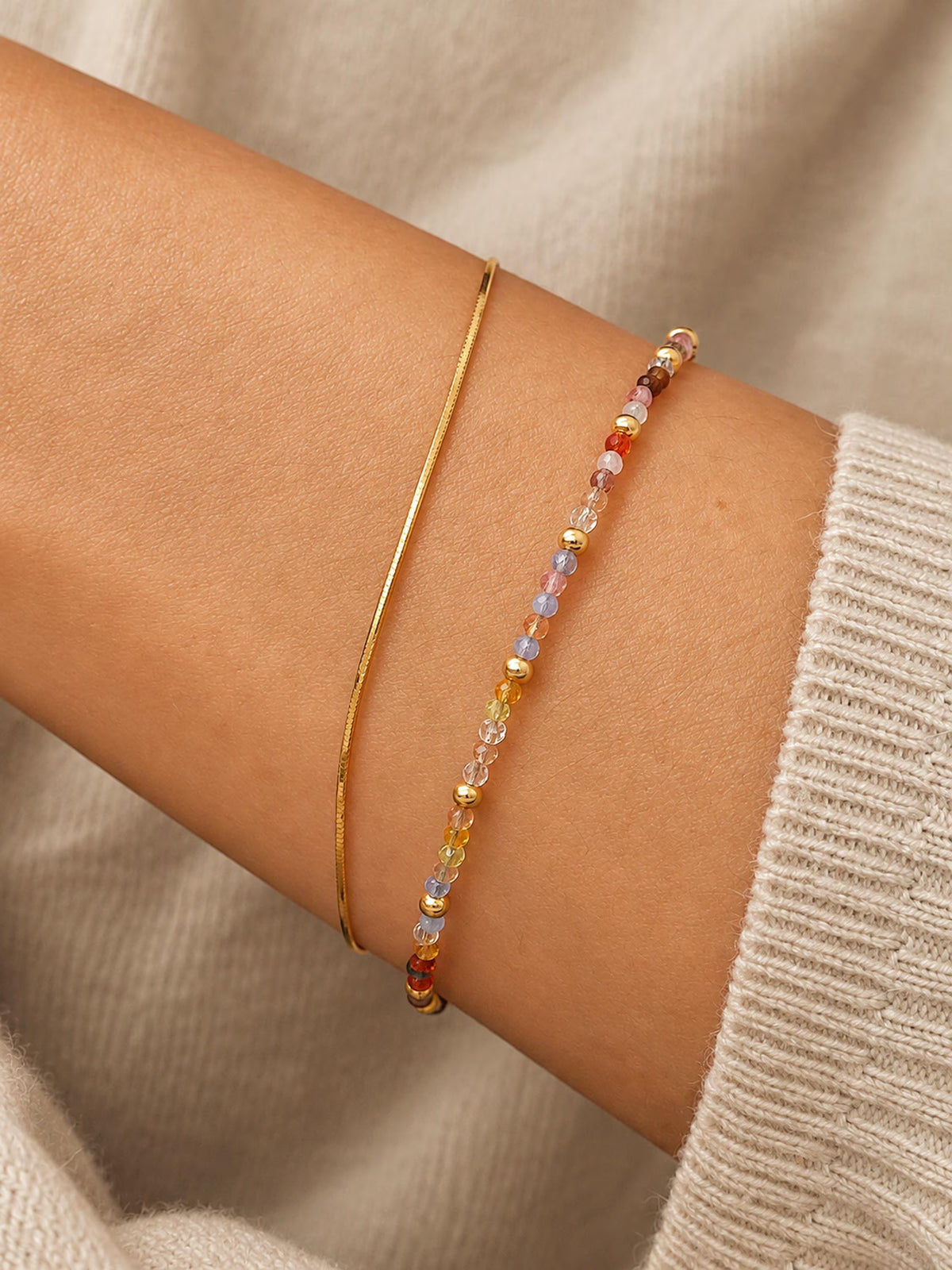 A close-up of a woman's wrist wearing two delicate bracelets: a thin gold bangle and a beaded bracelet with small multicolored faceted crystal beads.