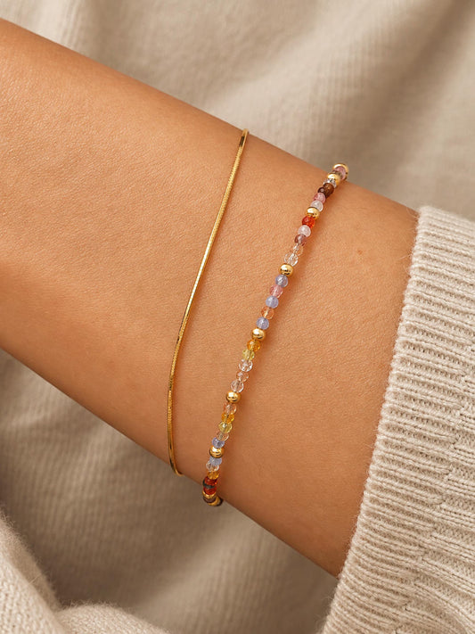 A close-up of a woman's wrist wearing two delicate bracelets: a thin gold bangle and a beaded bracelet with small multicolored faceted crystal beads.