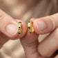 A close-up of two hands holding a pair of gold hoop earrings with small black and clear stones set along the front, against a soft beige background.