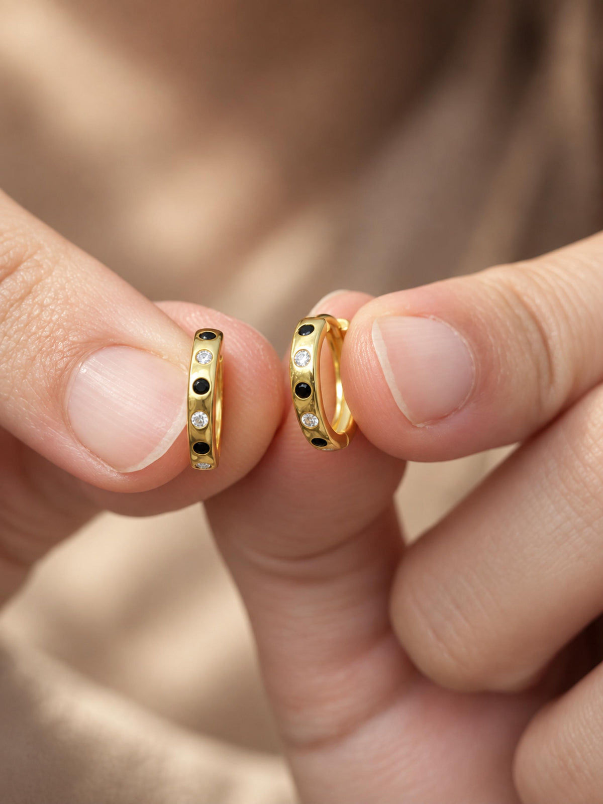 A close-up of two hands holding a pair of gold hoop earrings with small black and clear stones set along the front, against a soft beige background.