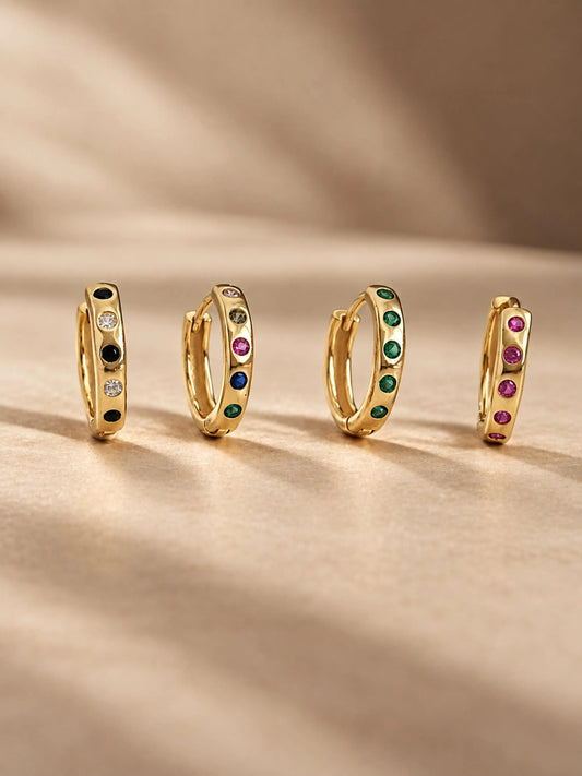 Four gold huggie hoop earrings with colorful gemstone accents, arranged in a row on a draped beige fabric background.