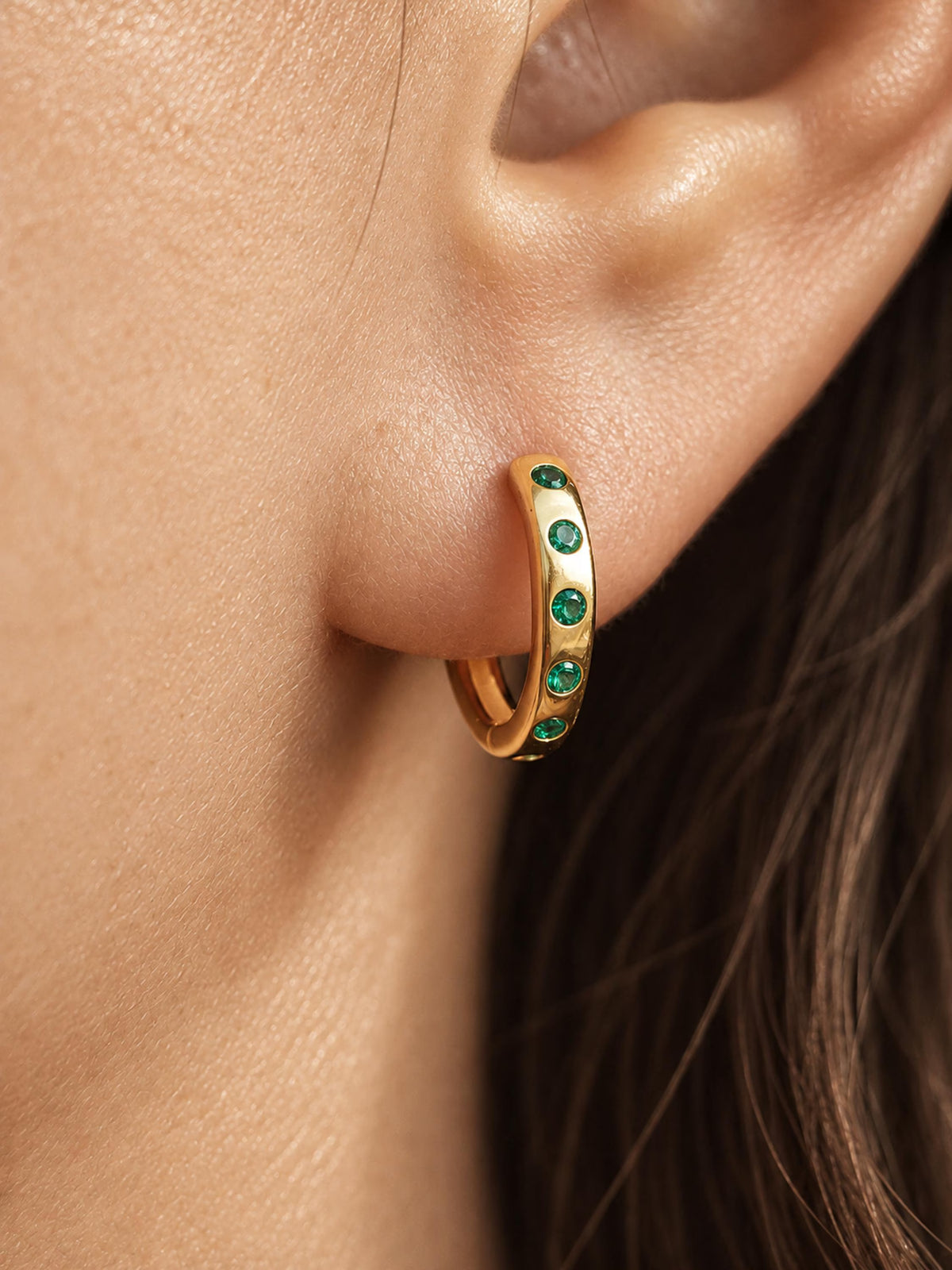 A close-up of a gold hoop earring adorned with small round green gemstones, worn on a woman's ear.