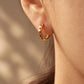A close-up of a person's ear wearing a small gold huggie hoop earring adorned with tiny red gemstones, set against a softly blurred warm beige background.