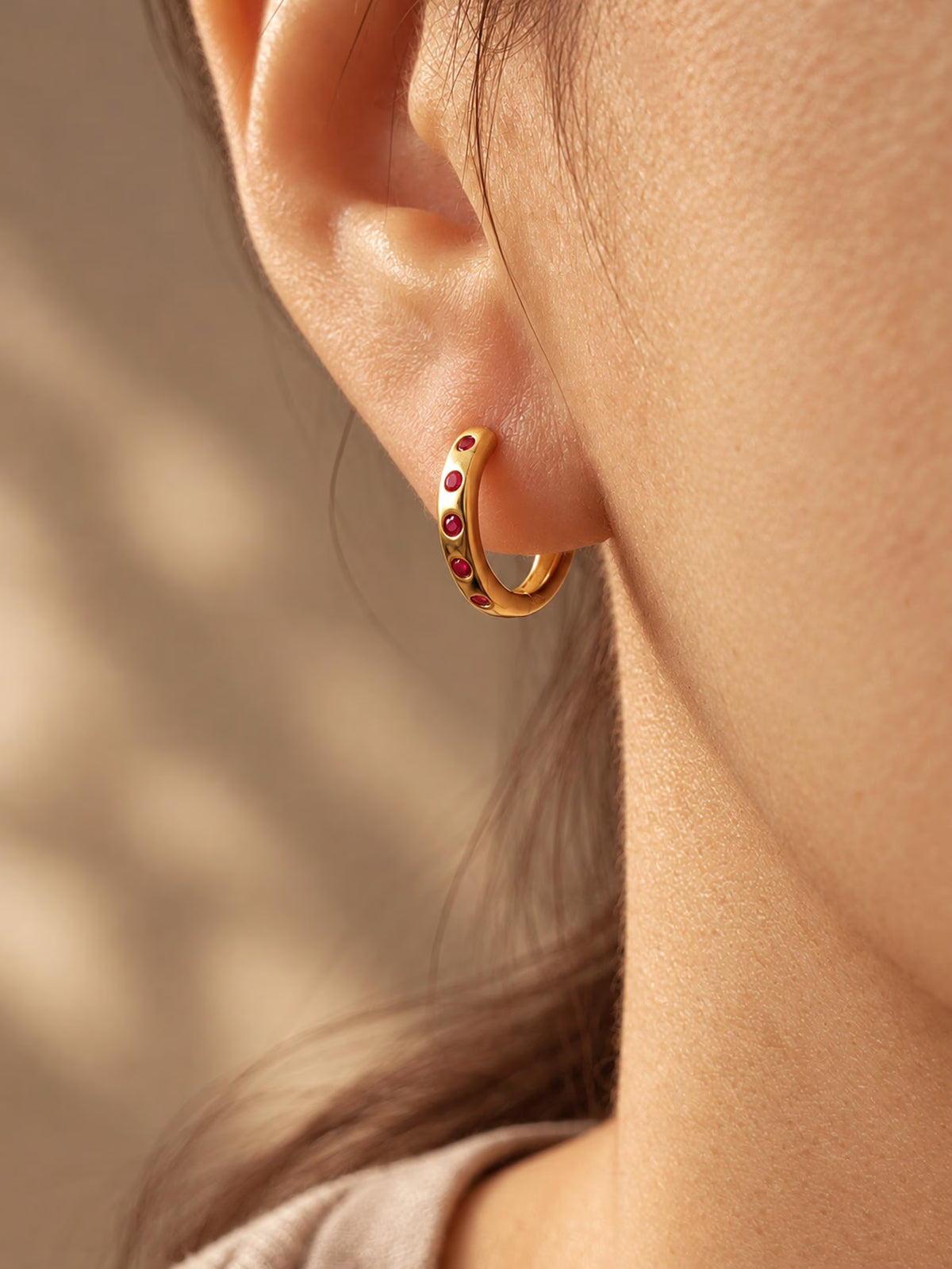 A close-up of a person's ear wearing a small gold huggie hoop earring adorned with tiny red gemstones, set against a softly blurred warm beige background.