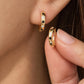 Close-up of a person's ear wearing a small gold hoop earring with alternating black and clear stones, while a hand holds a matching gold hoop earring against a skin-toned background.