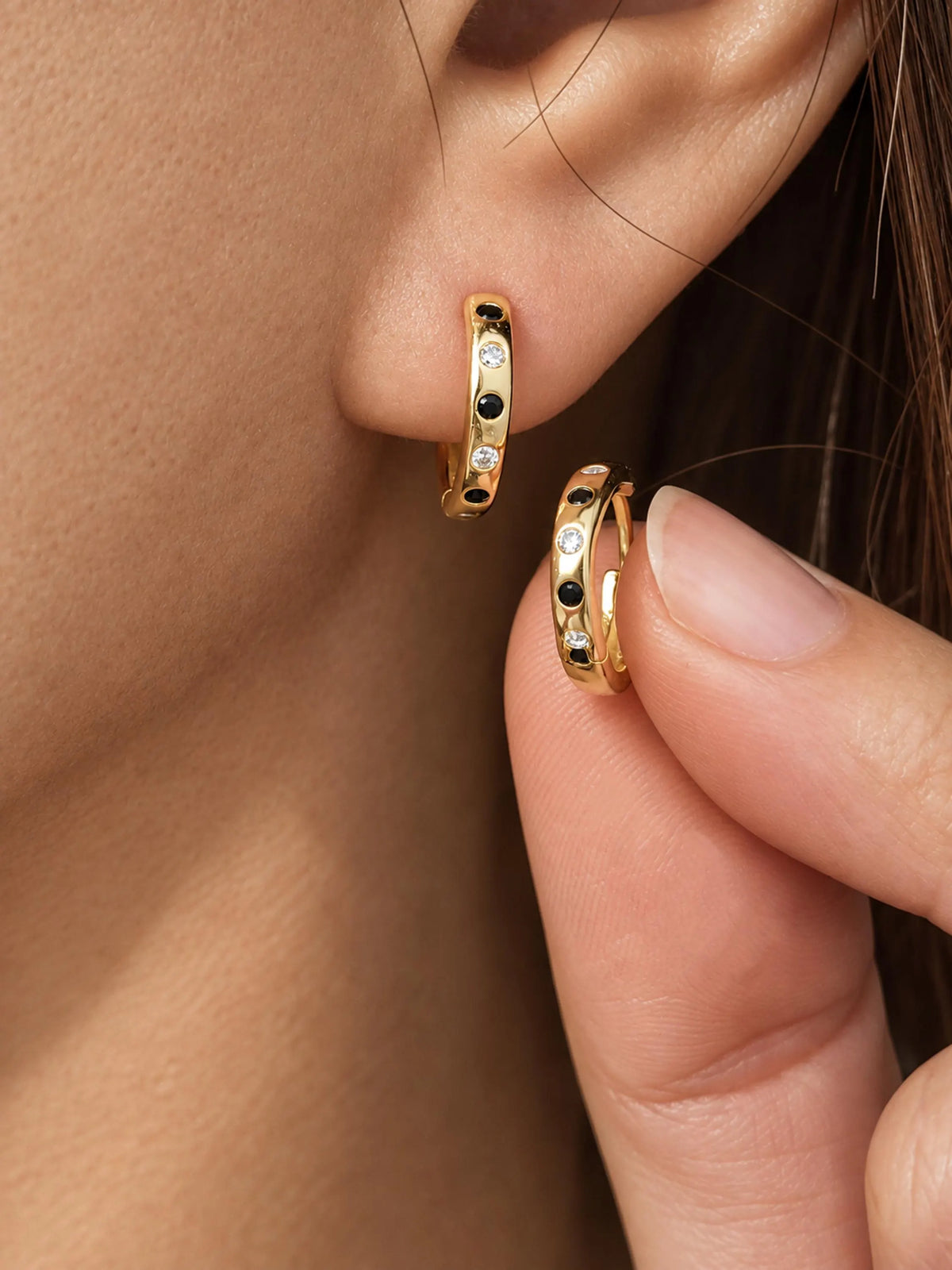 Close-up of a person's ear wearing a small gold hoop earring with alternating black and clear stones, while a hand holds a matching gold hoop earring against a skin-toned background.