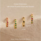 Four gold-plated huggie hoop earrings with cubic zirconia stones in pink, green, black, and multicolor settings, displayed on a textured beige background with text detailing the materials and dimensions.