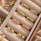 A close-up of multiple gold earrings displayed in a beige jewelry box. Each ring has small colored stones set along the band, including green, pink, blue, yellow, and black.