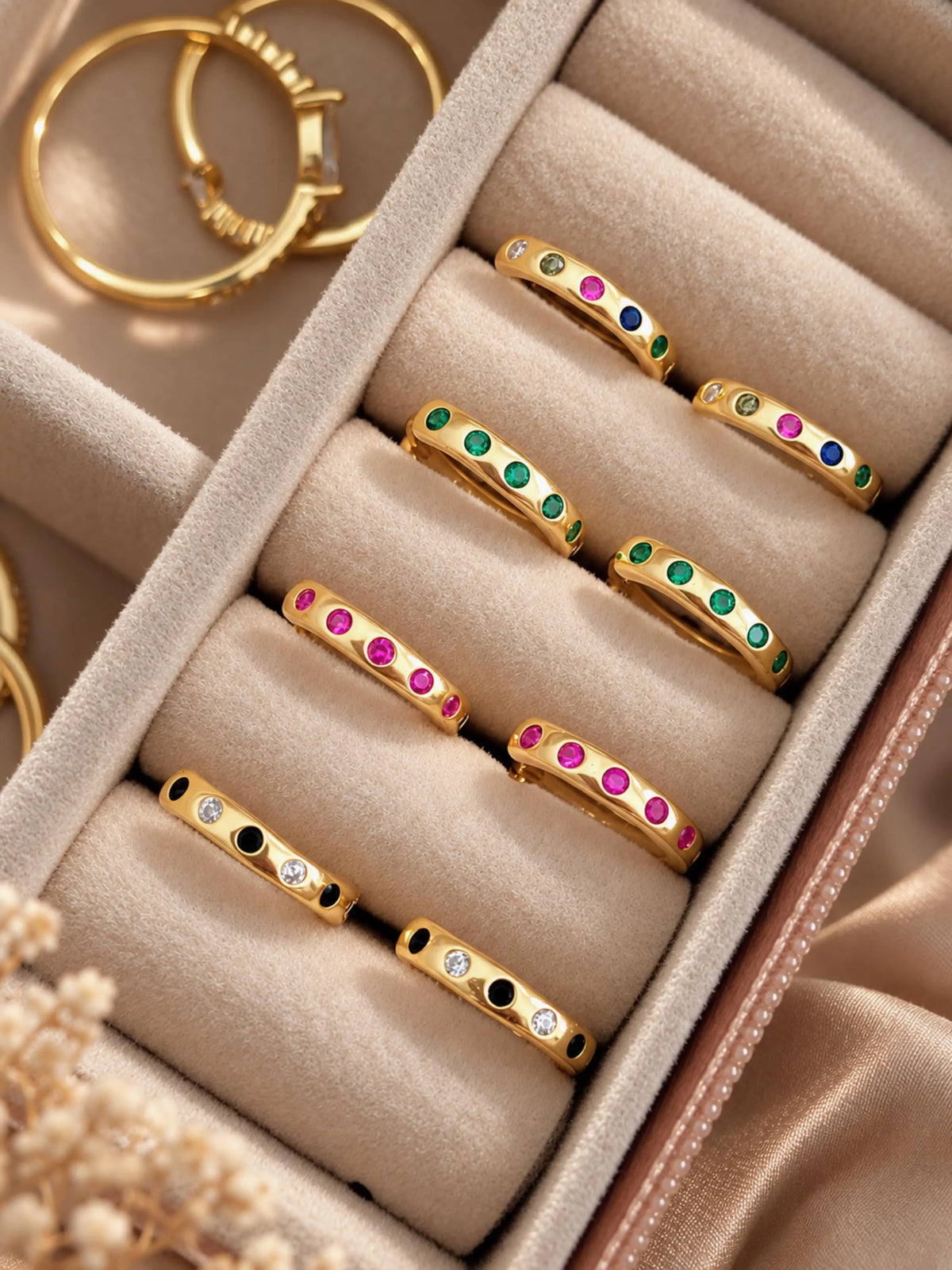 A close-up of multiple gold earrings displayed in a beige jewelry box. Each ring has small colored stones set along the band, including green, pink, blue, yellow, and black.