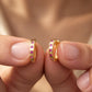 A close-up of two hands holding a pair of gold huggie hoop earrings with small pink gemstones set along the front, against a soft-focus warm skin-toned background.