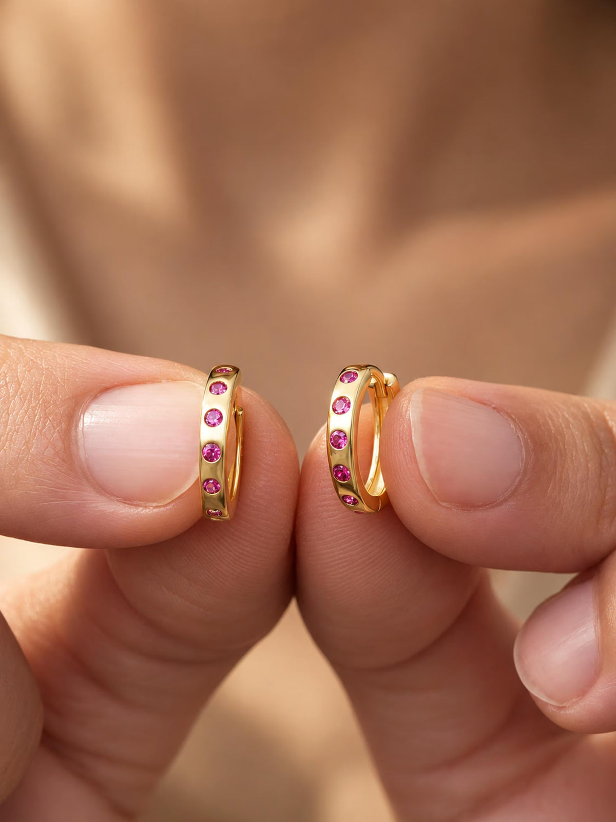 A close-up of two hands holding a pair of gold huggie hoop earrings with small pink gemstones set along the front, against a soft-focus warm skin-toned background.