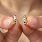 A close-up of a person's fingers holding a pair of small gold huggie hoop earrings adorned with colorful round gemstones in green, blue, pink, and purple.