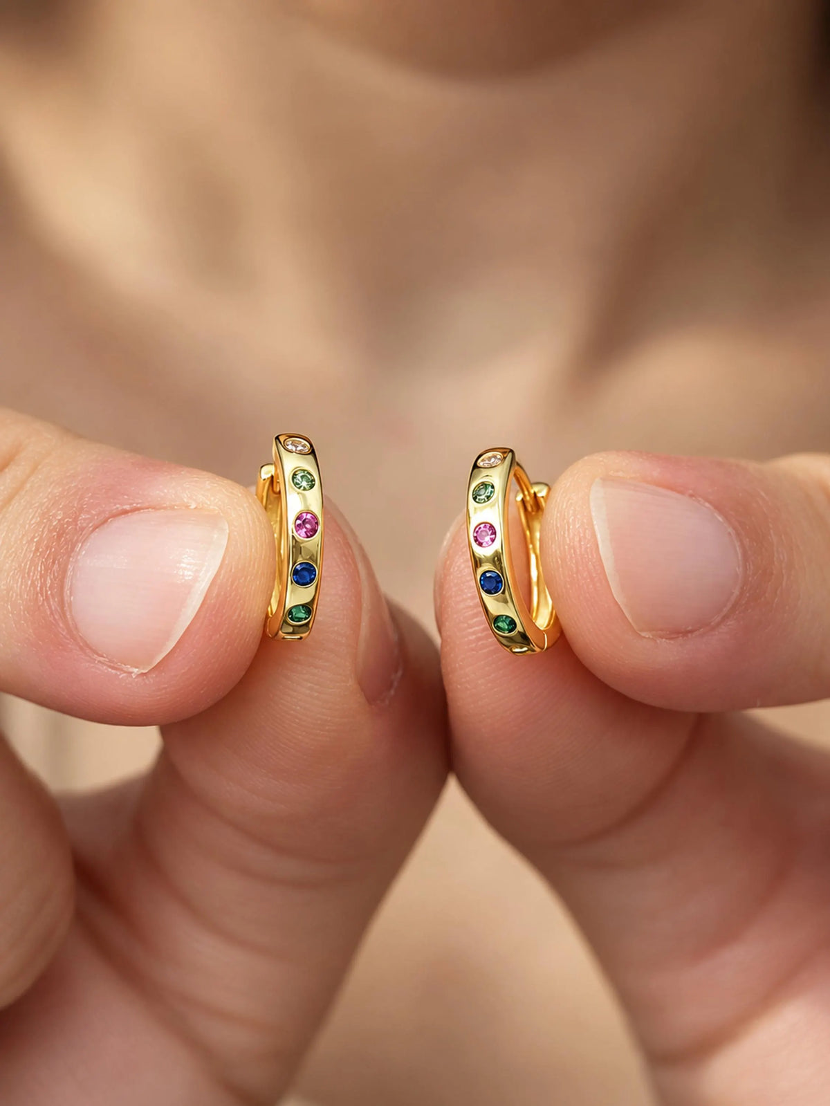 A close-up of a person's fingers holding a pair of small gold huggie hoop earrings adorned with colorful round gemstones in green, blue, pink, and purple.