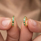 A close-up of a person's fingers holding a pair of gold hoop earrings adorned with small round green gemstones, set against a soft, blurred skin-toned background.