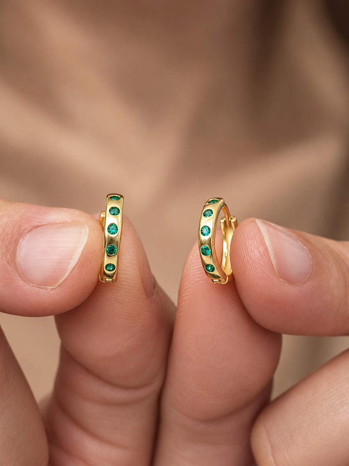 A close-up of a person's fingers holding a pair of gold hoop earrings adorned with small round green gemstones, set against a soft, blurred skin-toned background.
