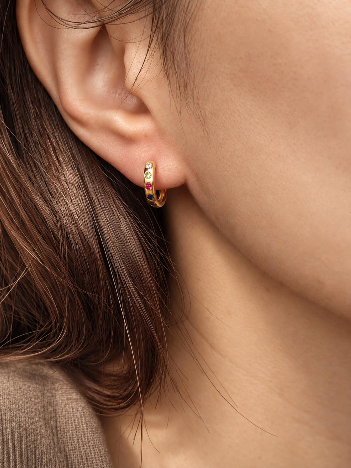 A close-up of a model's ear wearing a small gold huggie hoop earring adorned with tiny colorful gemstones.