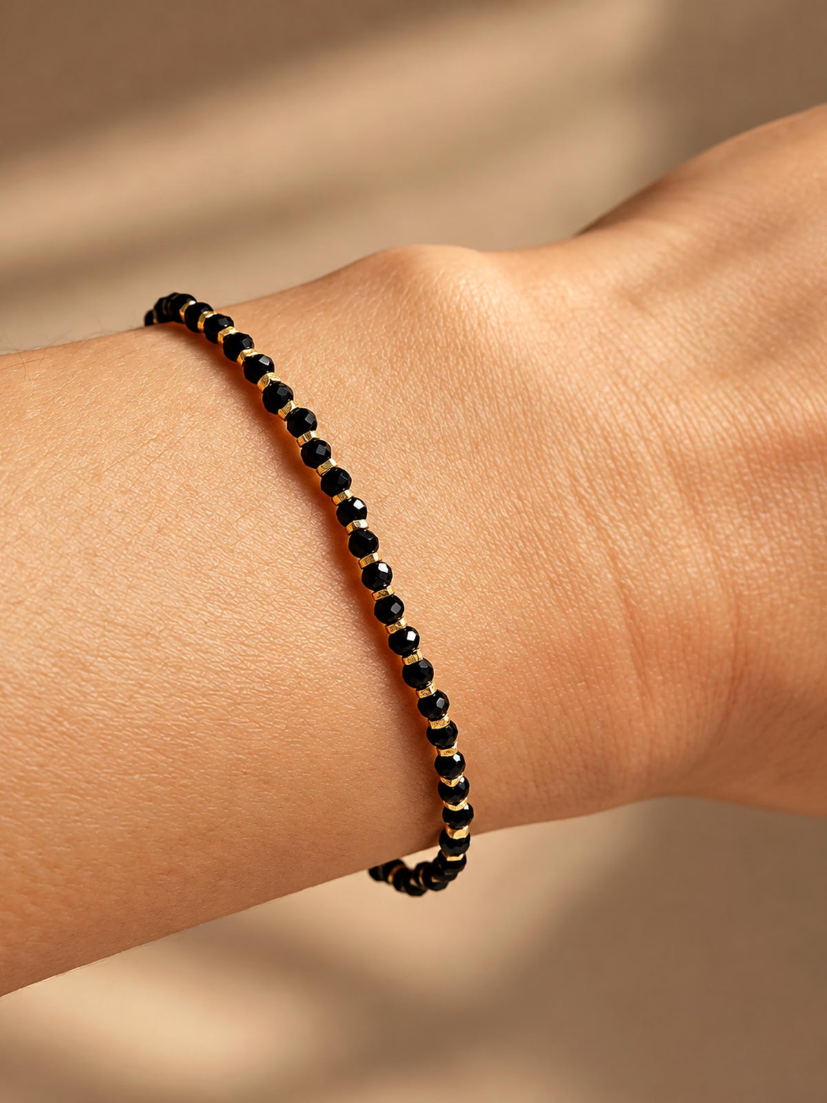 Close-up of a black beaded gemstone bracelet with small gold spacer beads worn on a woman's wrist against a warm beige background.