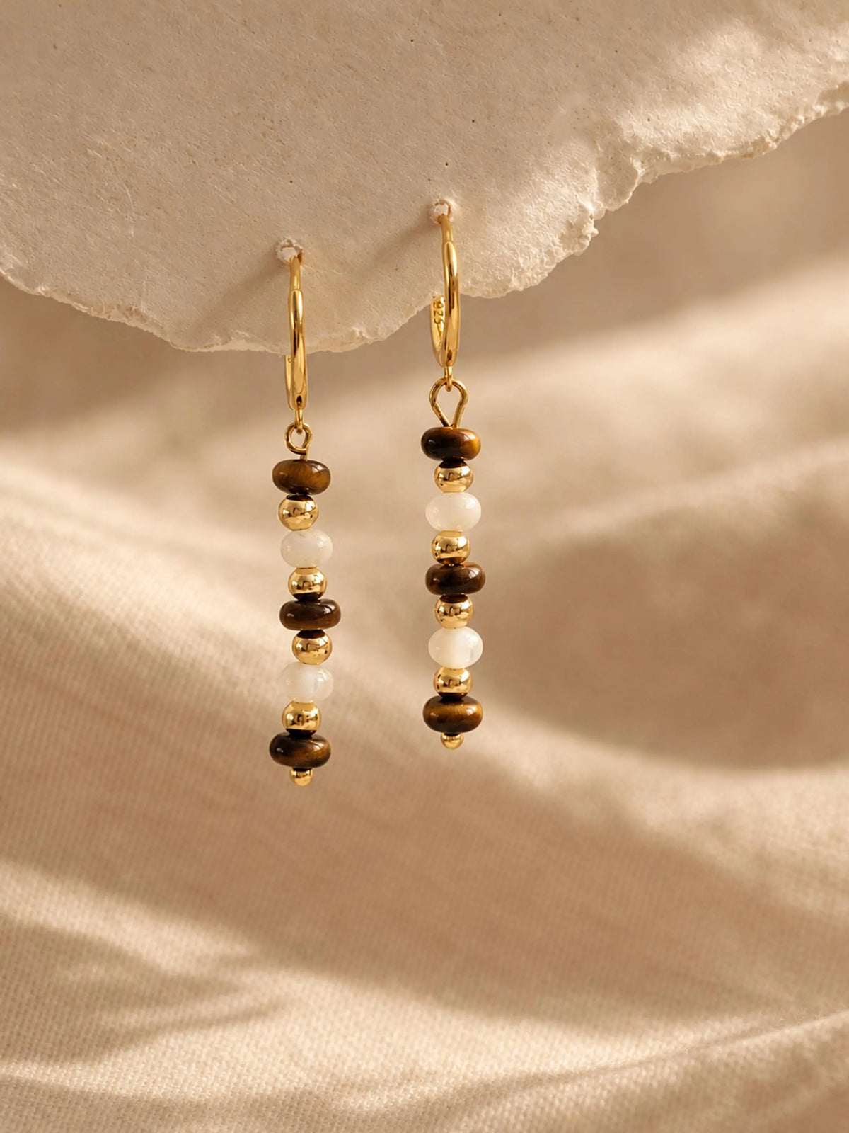Pair of Tiger's Eye and Mother of Pearl gemstone bar earrings dangling against a warm neutral background.